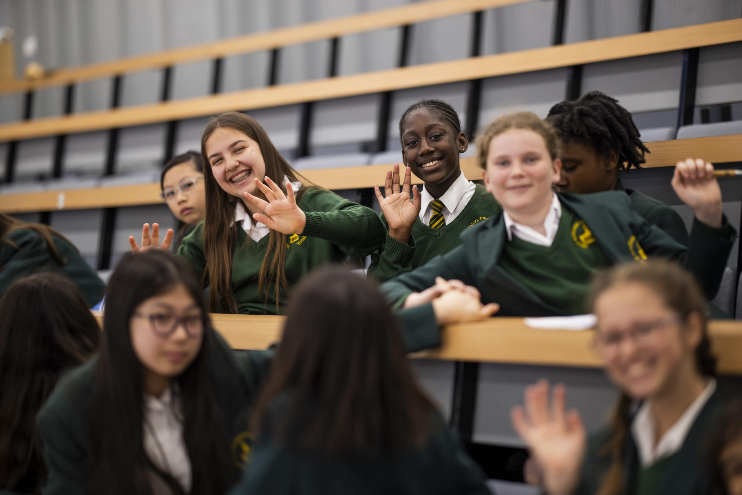 A group of smiling school children wearing green school uniforms sitting at modern style decks waving at the camera
