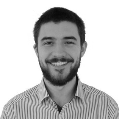 Black and white image of a fully bearded man smiling and facing the camera wearing a striped open collared shirt. Name is Andy Tomlinson