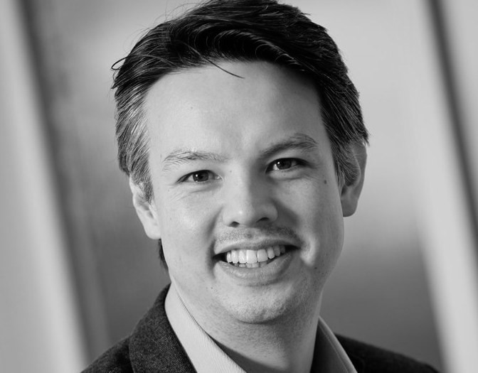 Black and white headshot of a man standing side profile smiling at the camera named Daniel Dyball