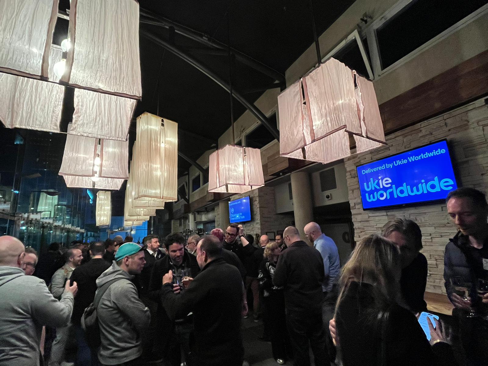 Crowd networking at a Ukie Worldwide event with ambient lighting and digital screens displaying the Ukie Worldwide logo