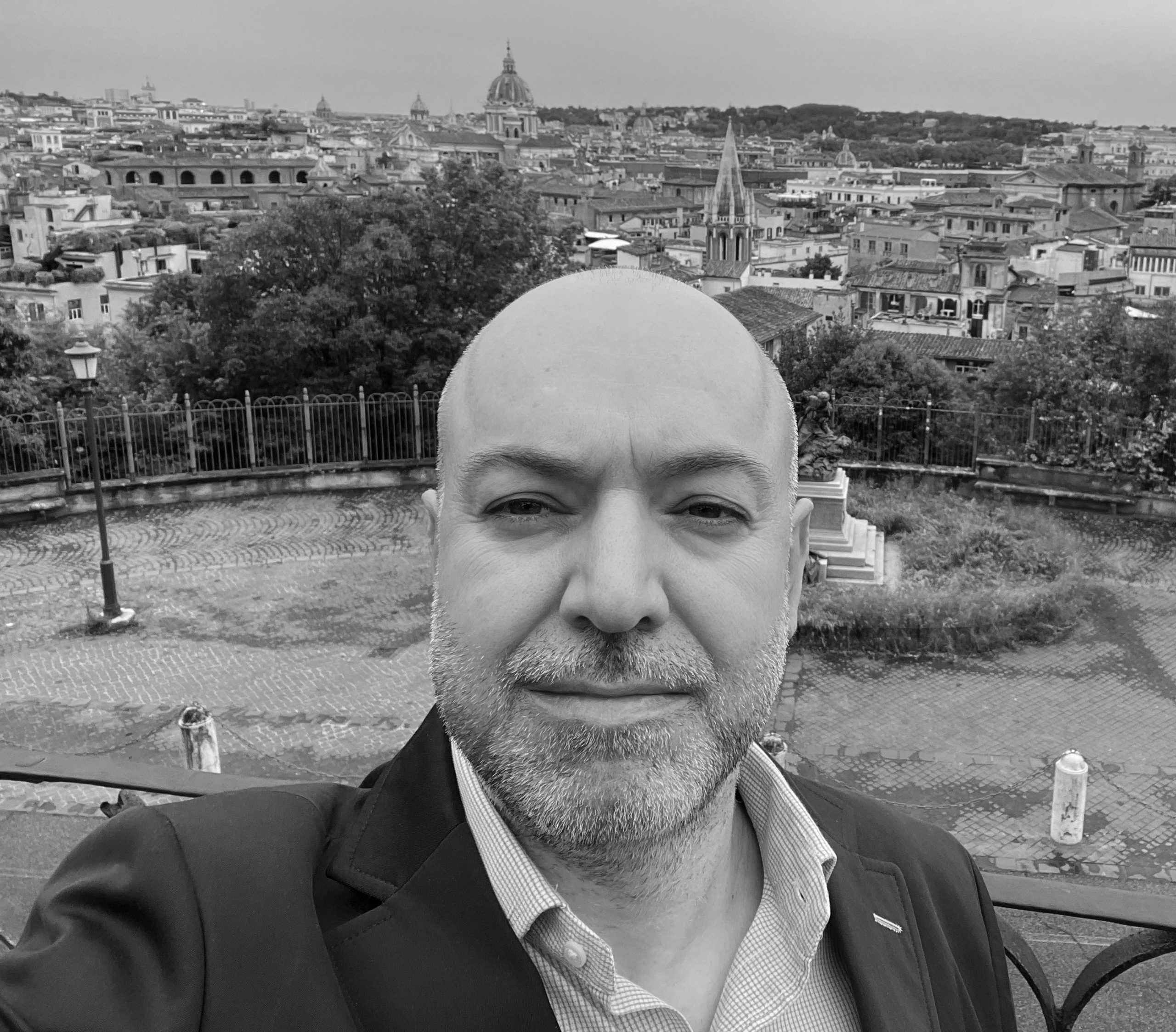 Black and white selfie image of a bald man with a beard standing in front of a cityscape. His name is Gustavo Herrera