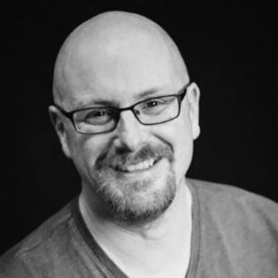 Black and white image of a bald, bearded man who is wearing glasses smiling at the camera. His name is Nick Button-Brown