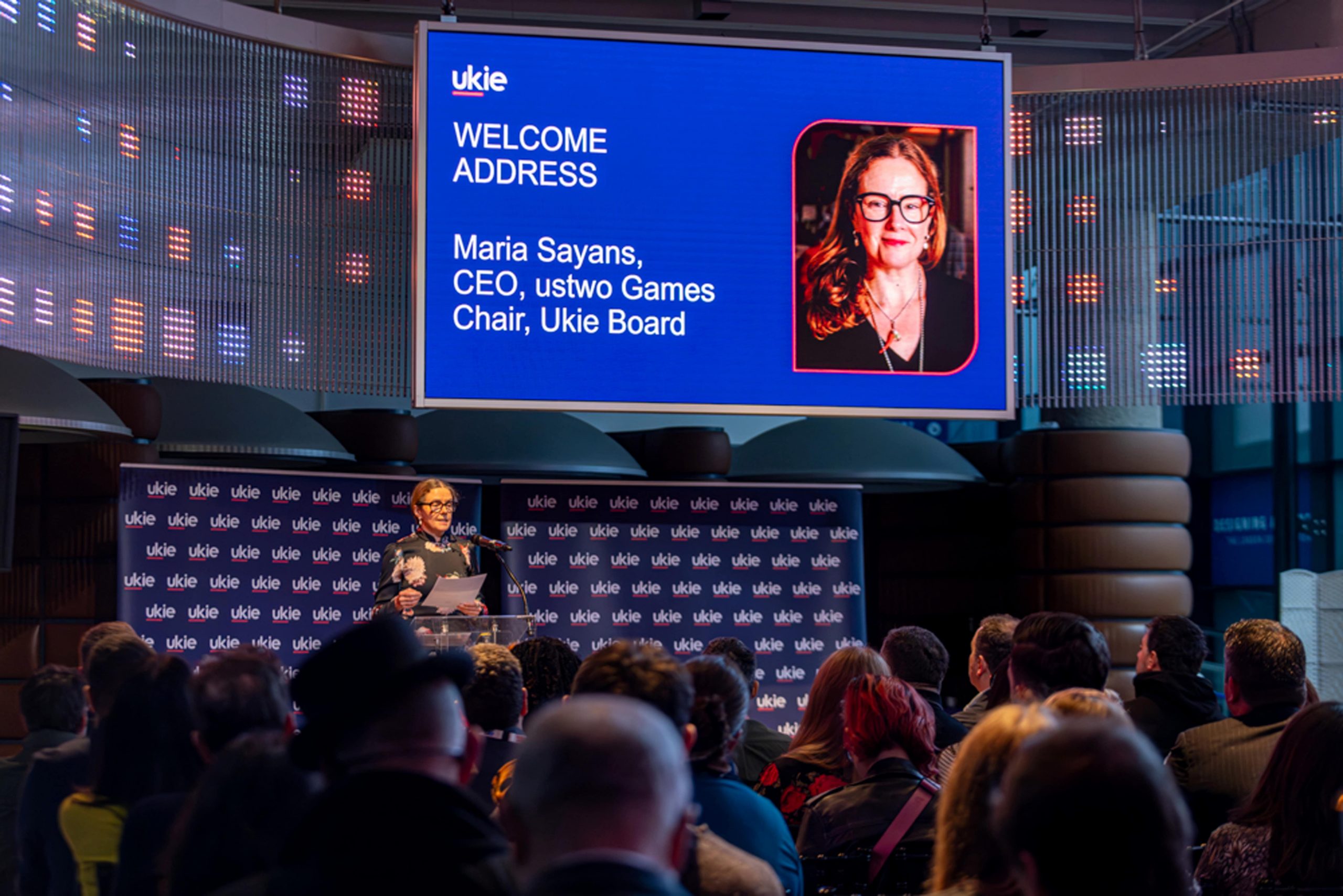 Professional women standing at a lectern giving a speech in front of a crowd, with a blue display in the background with the Ukie logo and her image with text