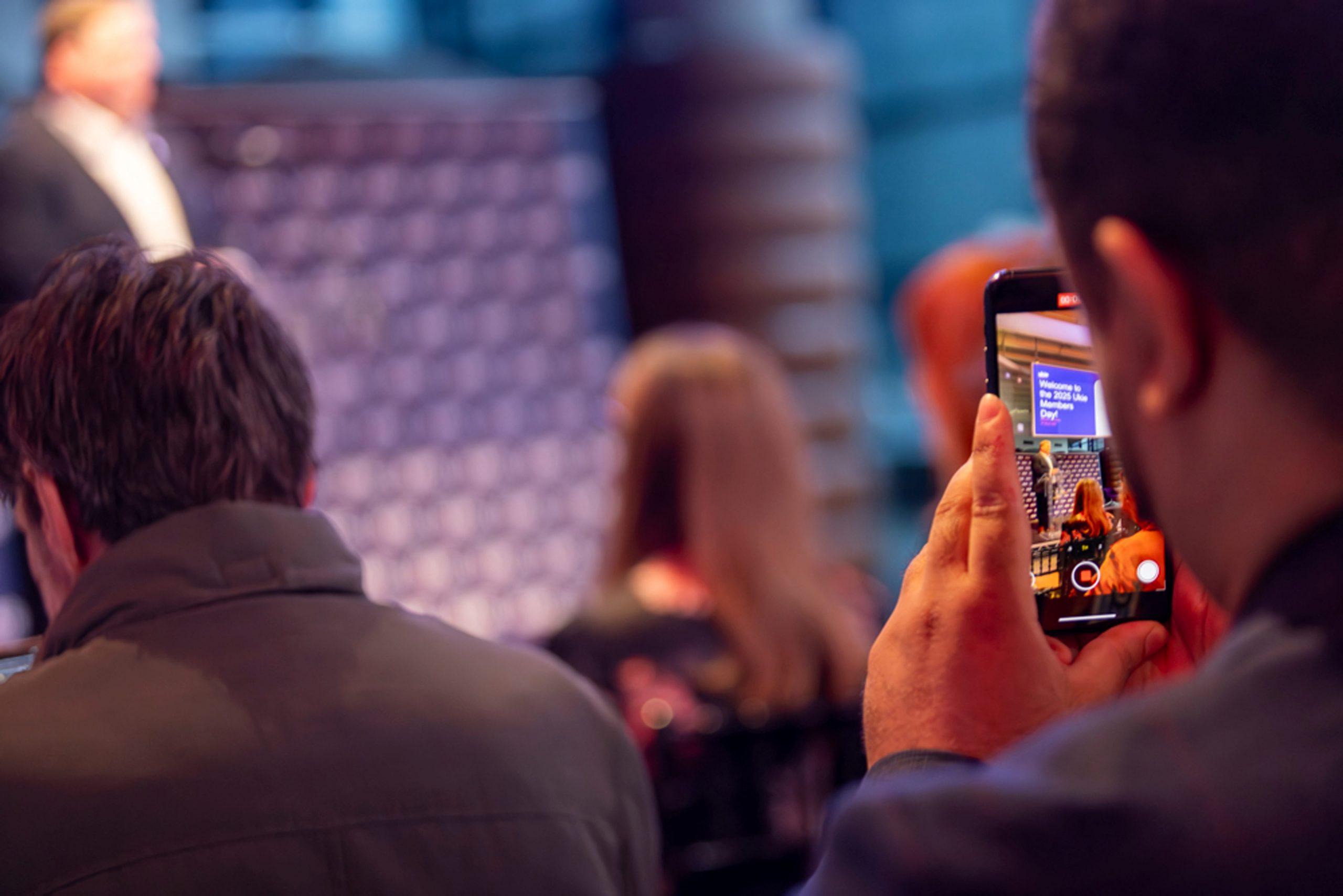 Close up of man holding a phone recording a speech by a professional on a stage at Ukie members day
