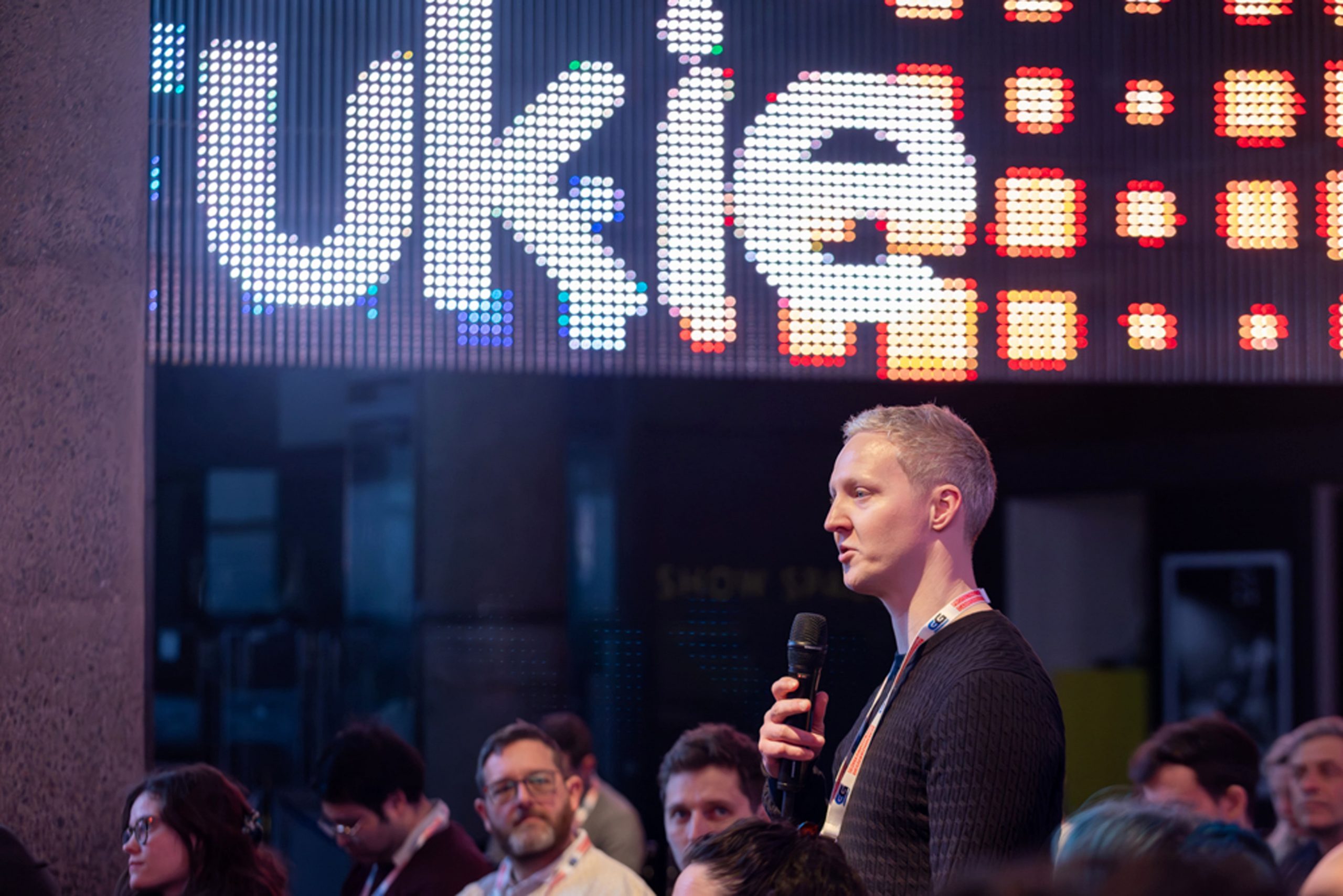 Man standing up at a professional event with a microphone asking a question in a crowd. Large digital sign in background displaying 'Ukie' with illuminous pattern