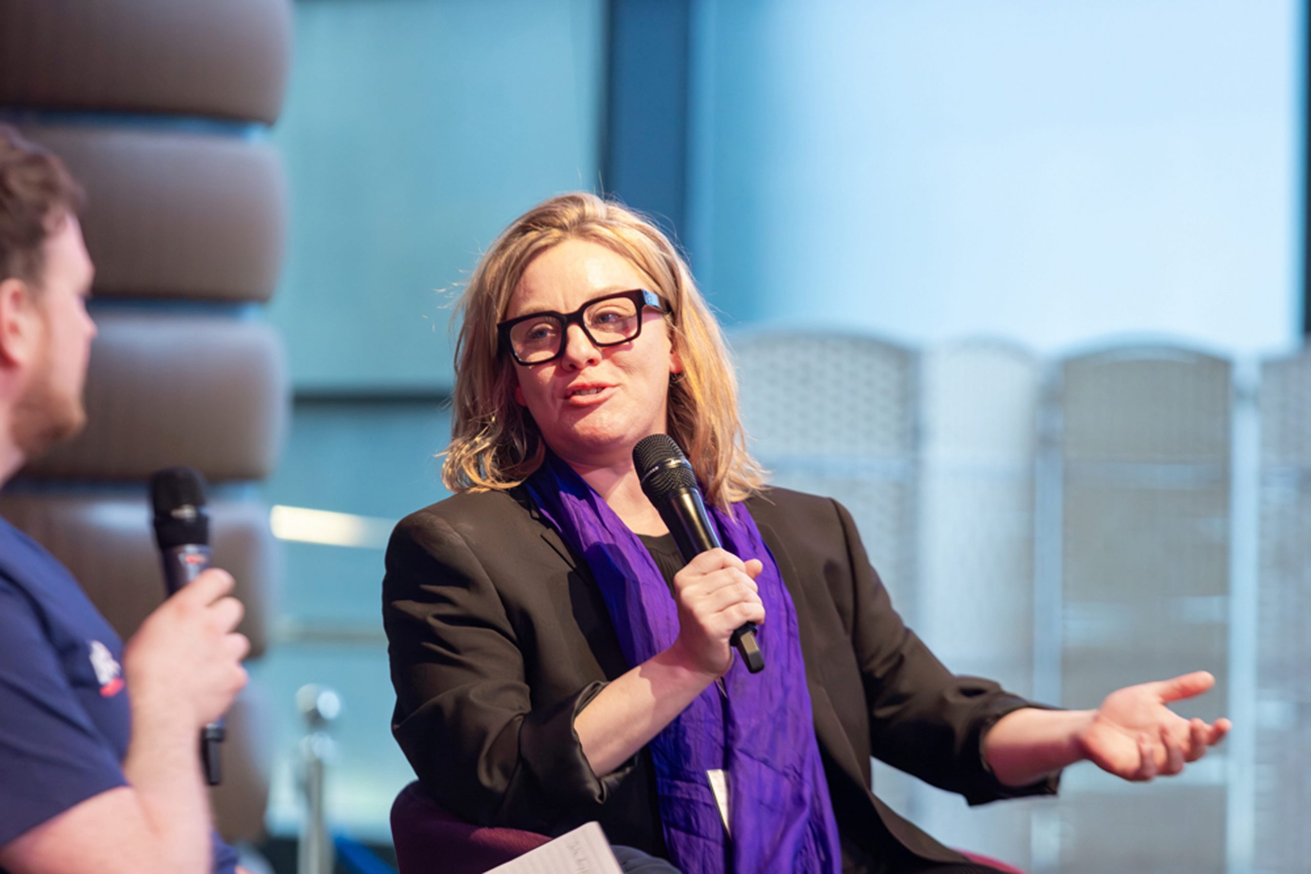 Professional looking women wearing glasses and a purple scarf taking part in a discussion with a microphone