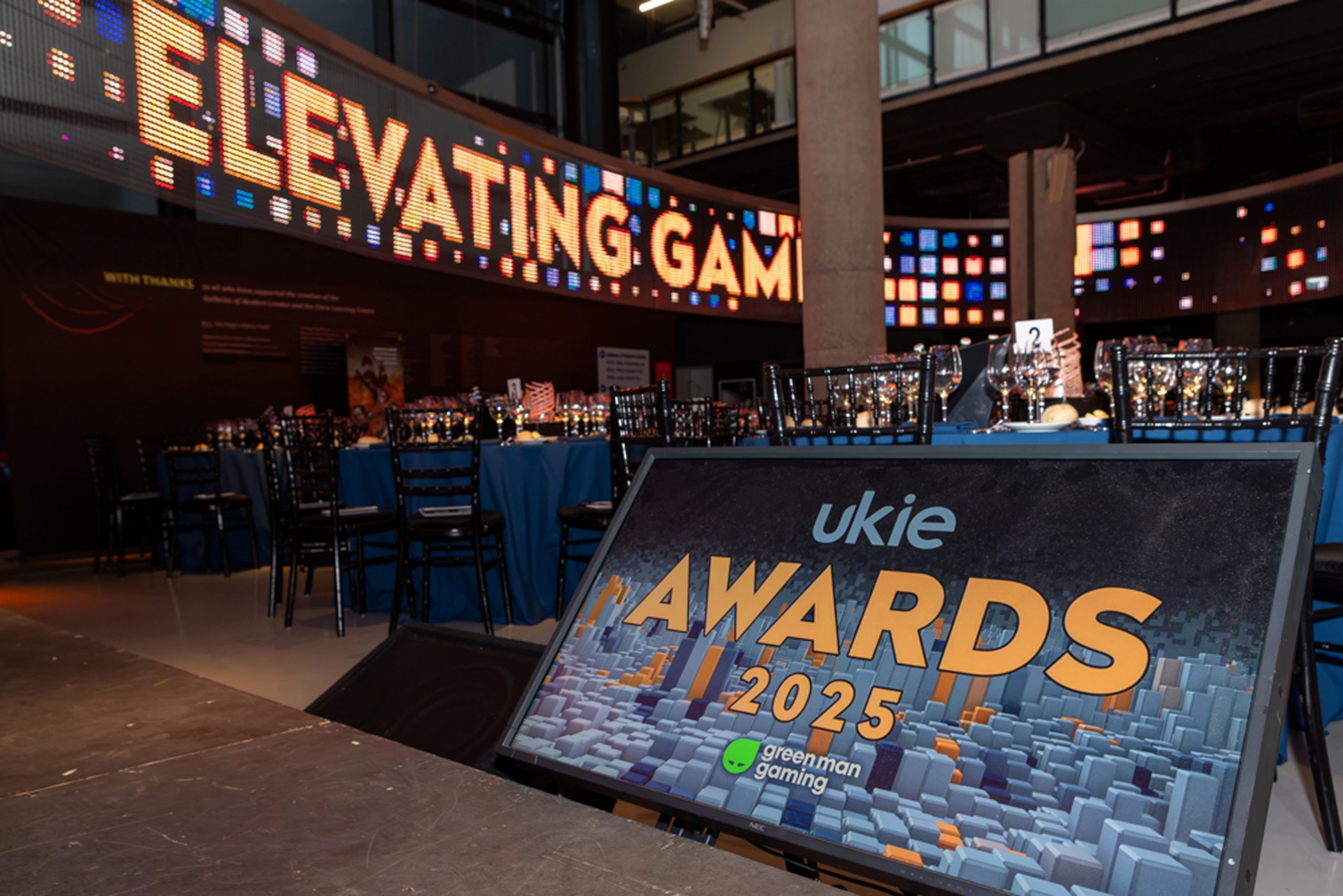Ukie Awards 2025 event setup with banquet tables, chairs, and a digital sign displaying 'Elevating Gaming' in the background and an 'Ukie Awards 2025' display board in the foreground sponsored by Green Man Gaming