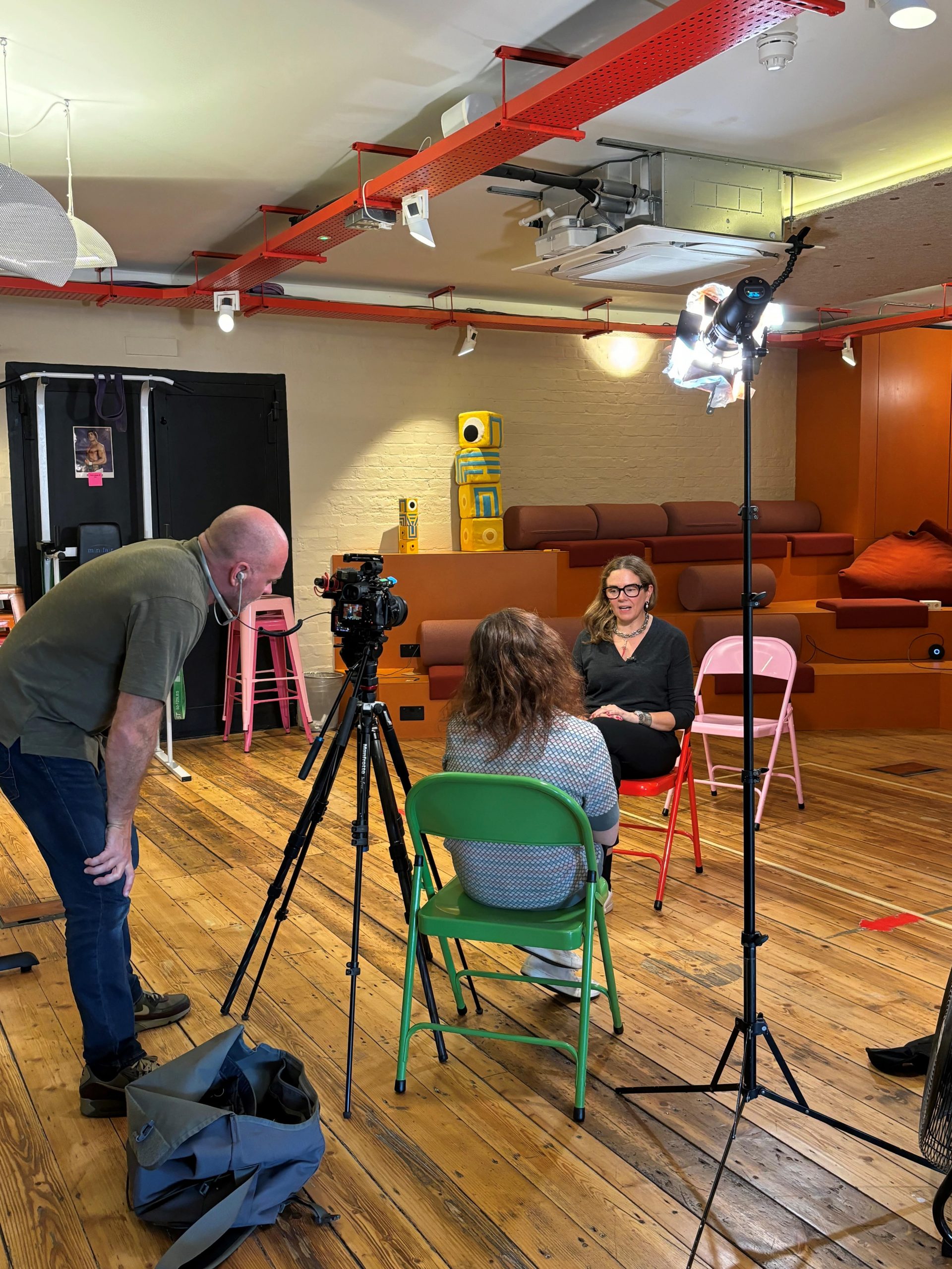 Behind-the-scenes setup of an interview with a woman seated under studio lights, being filmed in a modern, casual workspace