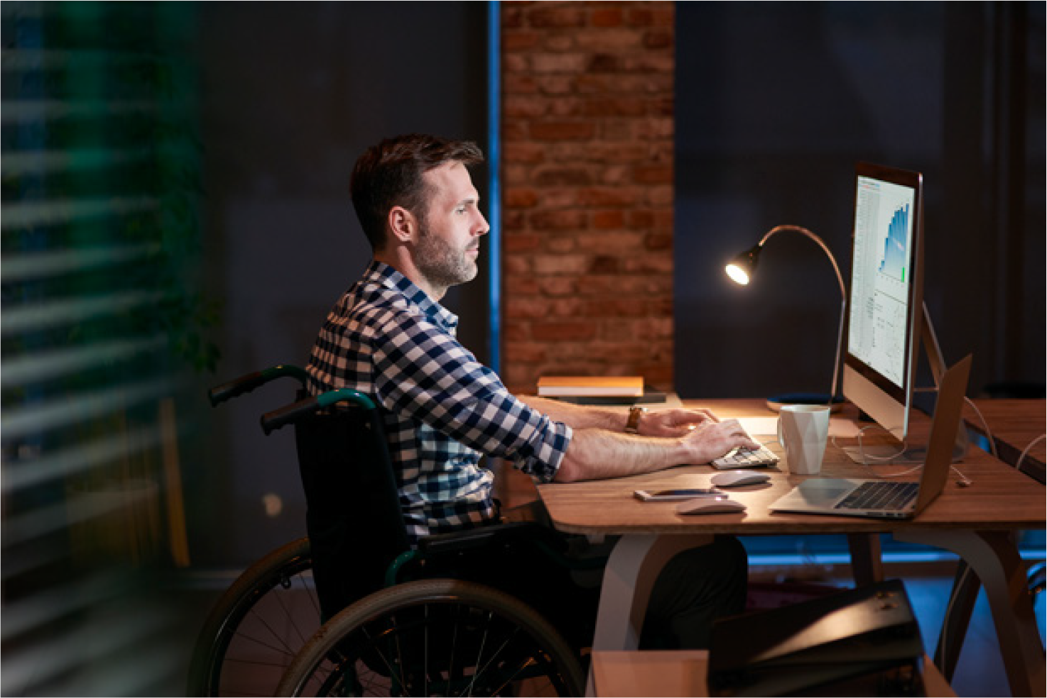Man in a wheelchair working at a desk with multiple computer screens in a modern office setting at night