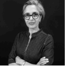 Black and white portrait of a woman with short hair and glasses, wearing a dark top, standing with arms crossed against a plain background. Her name is Reedah El-Saie